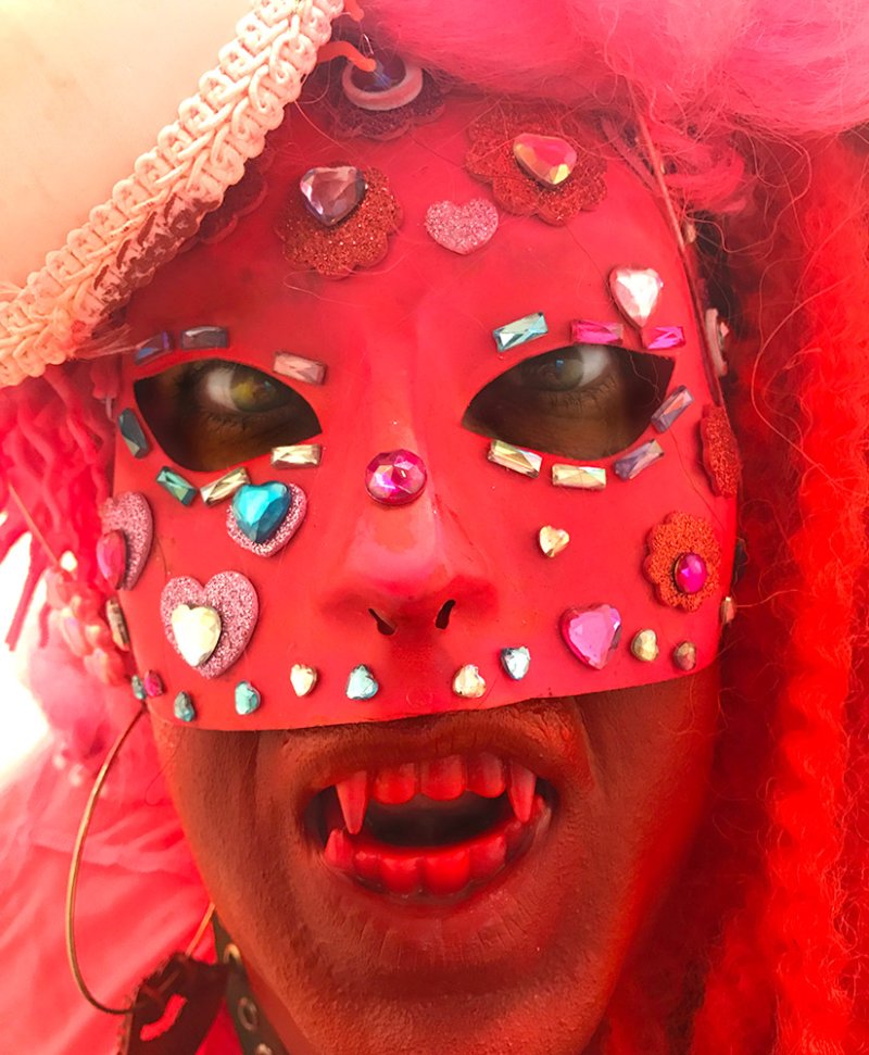 Close-up of a person in a red mask adorned with glittery hearts and gems, red dreadlocks, and vampire fangs.
