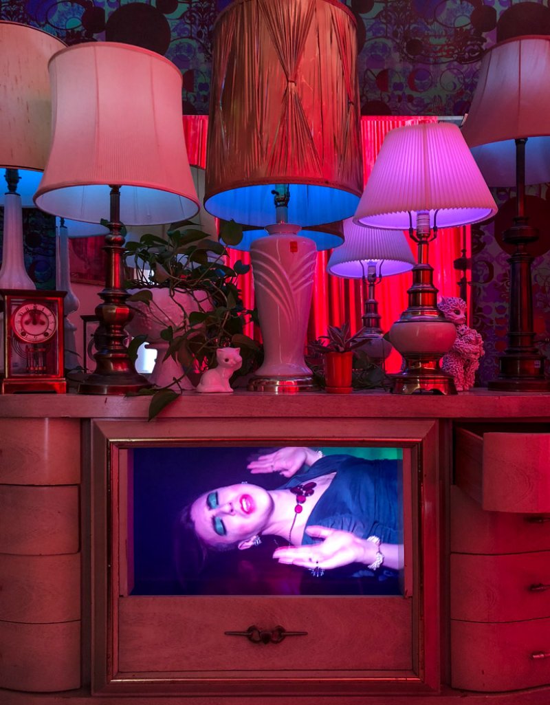 A woman with dark hair and bright makeup is displayed on a screen within a dresser, surrounded by lamps.
