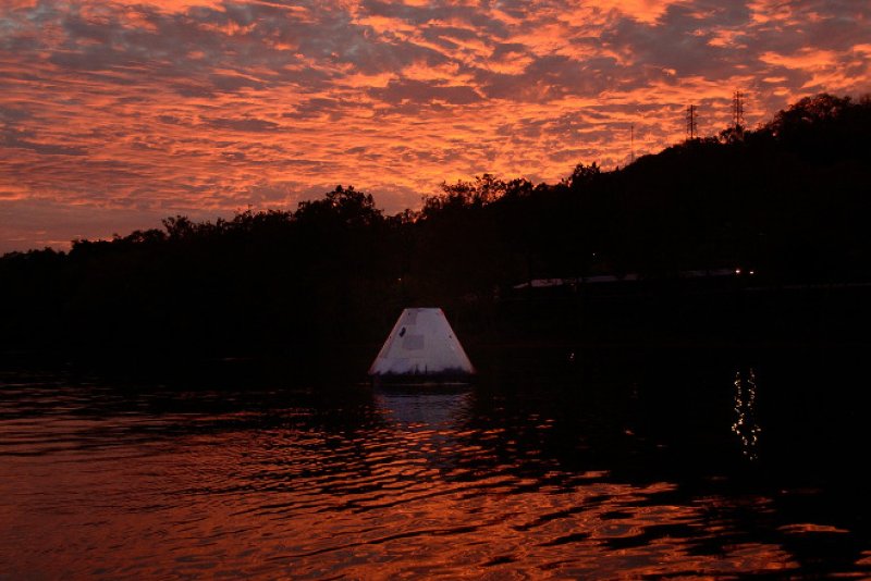 Artwork depicts a white, cone-shaped object in water, reflecting a fiery orange sky. A dark, silhouetted treeline and hill form the background. The water's surface is rippled with orange light.