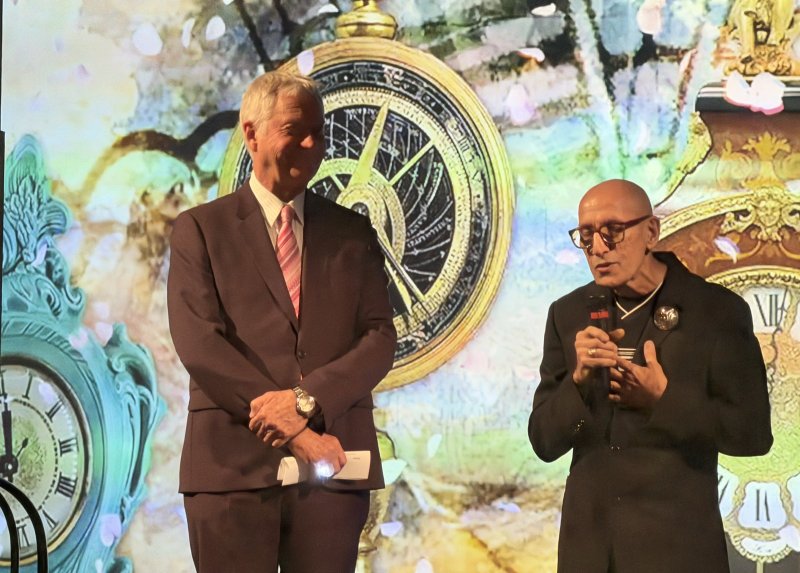 Two men on stage, one in a suit, one in black with glasses, against a backdrop of clocks and falling petals.