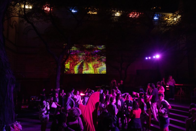 Night scene of a crowd under trees, bathed in purple light. A colorful abstract projection is visible on a building behind the crowd, with lights above.