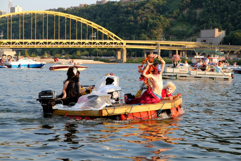 Three costumed people on a small, makeshift boat with an outboard motor, on a river with a yellow bridge in the background.