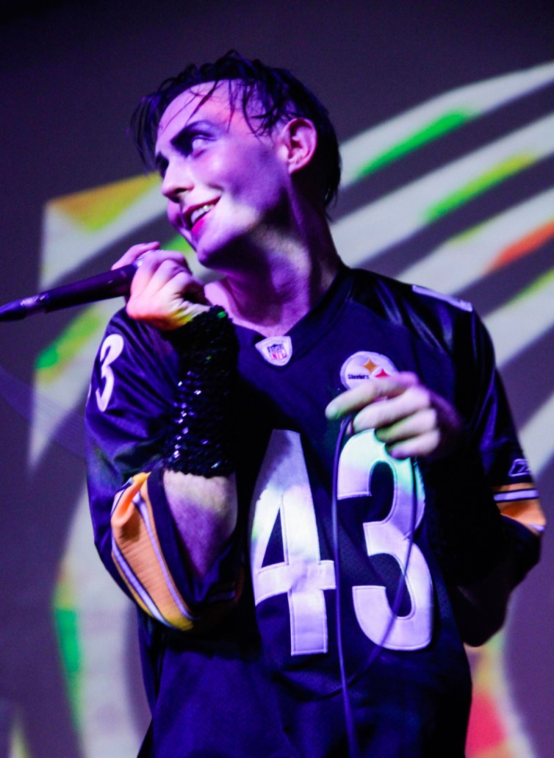 Performer in Steelers jersey, sequined sleeve, holding microphone under purple lighting with striped background.