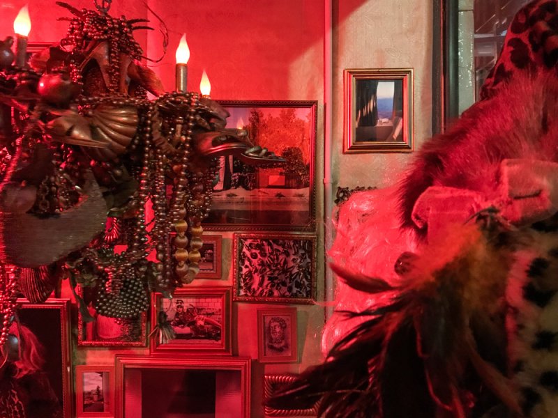 A dark room bathed in red light, featuring a chandelier with flickering candle-like bulbs, framed artworks, and taxidermy.