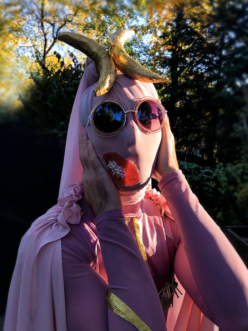 Person in pink full-body suit, round sunglasses, and red beaded lips, holding face, with golden bananas on head.