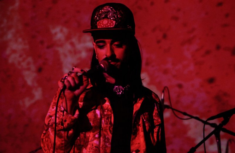 Red-toned portrait of a person singing into a microphone. They wear a patterned shirt, bejeweled cap, and necklace. A textured red background is behind them.
