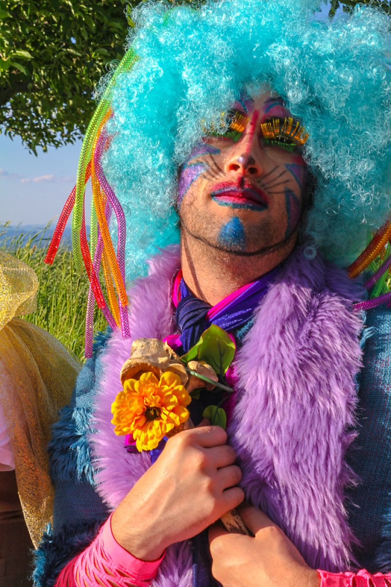 Person in a bright blue wig, colorful makeup, and a purple faux fur vest holds a yellow flower.
