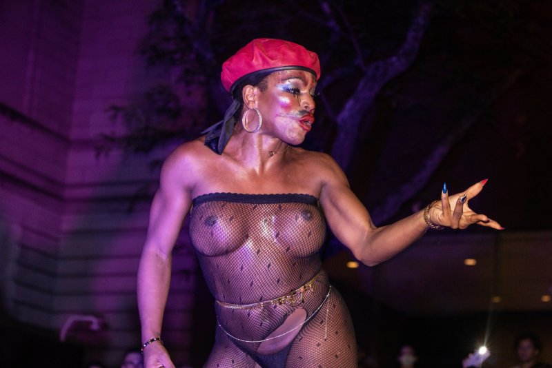 Performer in fishnet bodysuit and red beret poses under purple light. Bold makeup, hoop earrings, and gold chains accent the figure.