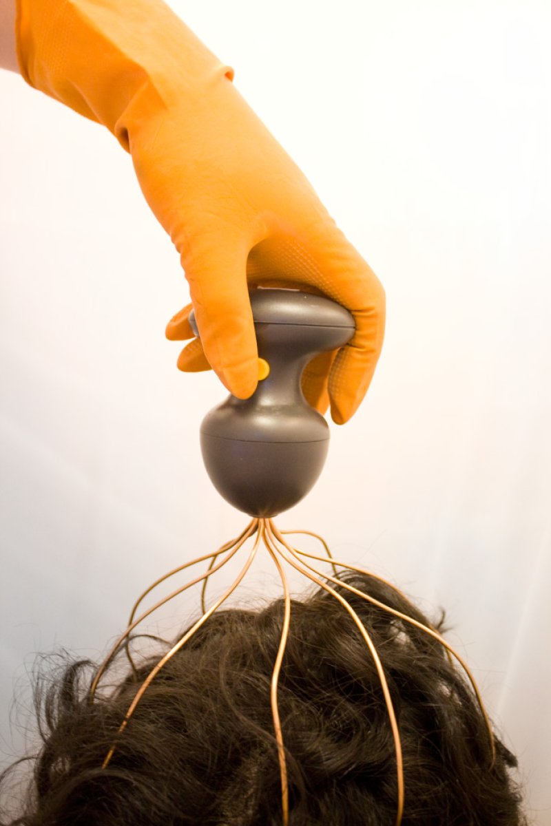 An orange-gloved hand holds a dark head massager with copper wires on a person's dark, curly hair.