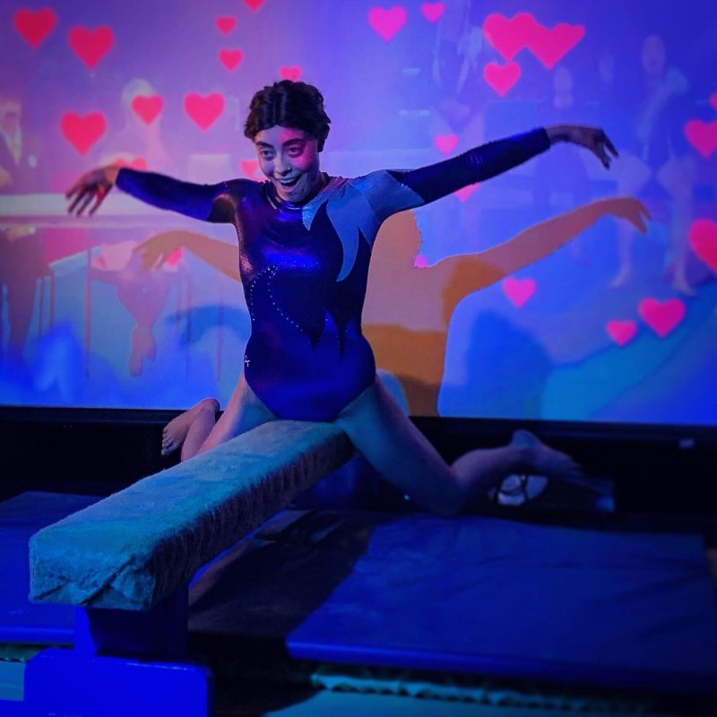Gymnast in blue leotard on balance beam, arms outstretched, pink hearts projected on blue background.