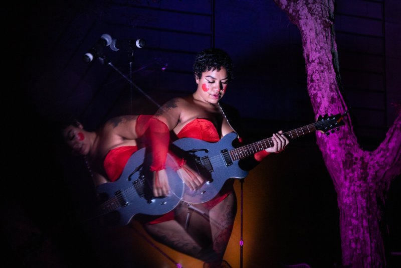 Performer in red corset and hosiery plays a blue electric guitar, with blurred ghosted image showing movement. Stage lighting is purple and white.