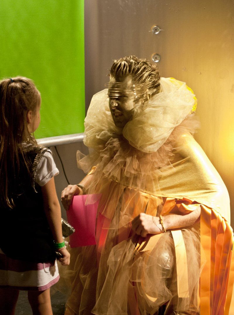 Gilded figure in ruffled gold costume and horizontal blindfold sits before a green screen, facing a young girl. Figure holds pink paper; metallic wall in background.