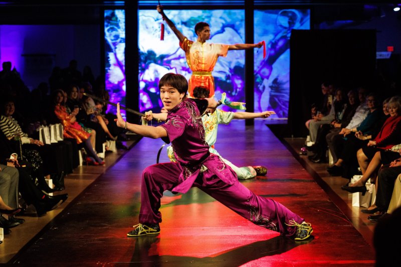 Three performers in colorful martial arts attire pose on a runway before an audience, with blue-lit screens behind them.