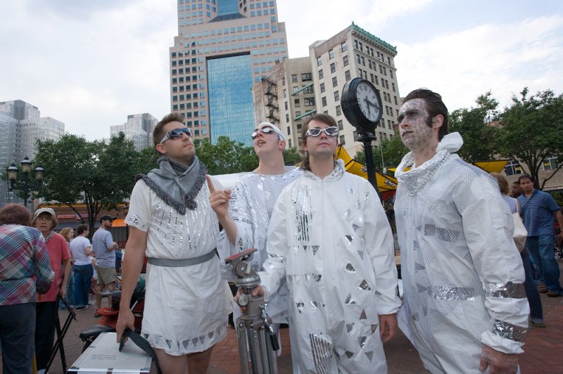 Four men in futuristic white and silver costumes and sunglasses stand outdoors in a city, looking up.