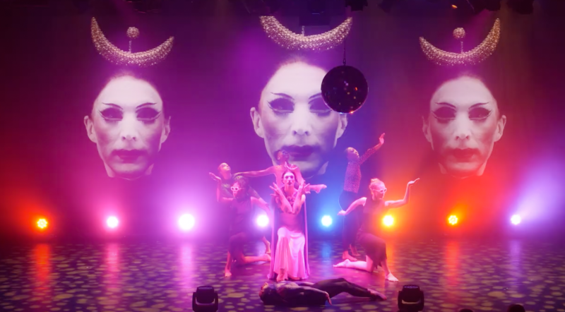 A stage performance with dancers in various poses under three large projections of a face with crescent moons, bathed in pink and purple light.