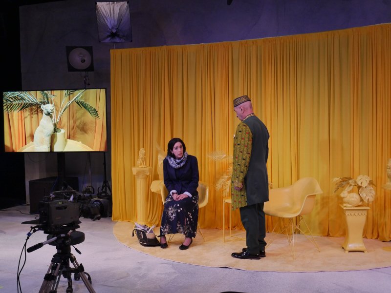 A woman in a dark dress sits on a chair while a man in a green and gold tunic stands facing her, all against a yellow backdro