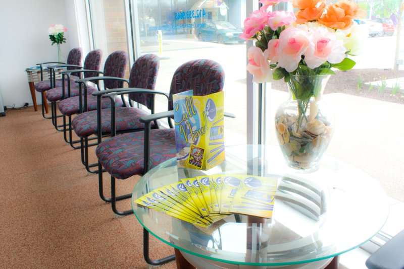 Waiting room with a glass table holding brochures and a vase of flowers, next to a row of chairs.