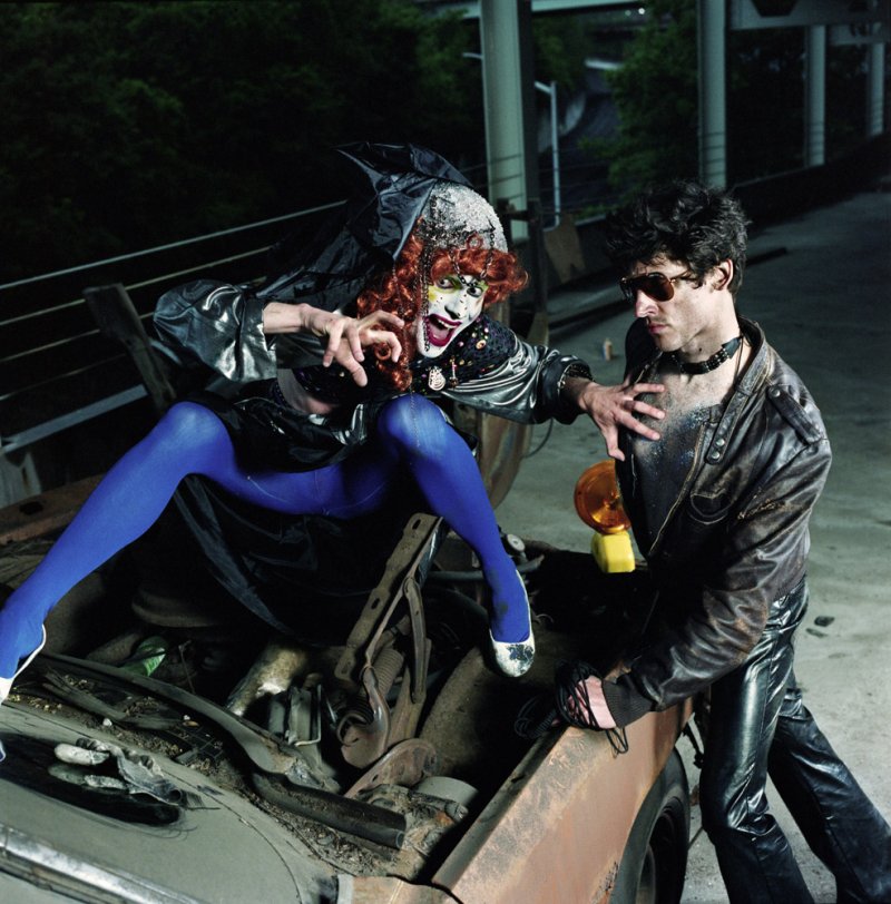 Drag queen in blue tights and witch hat lunges from car hood at man in leather jacket and sunglasses.