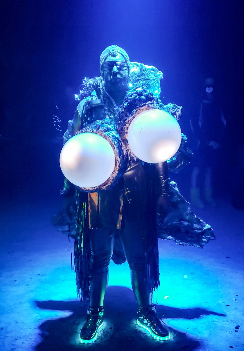 Performer in dark blue light, wearing a gold costume with two large glowing white orbs on the chest and light-up shoes.