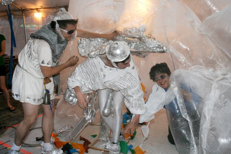 Three figures in white and silver costumes interact with a large, clear plastic structure. One figure wears a helmet and tunic, another is crouched, and the third is partially obscured by the plastic. Foil accents and playful details are visible.