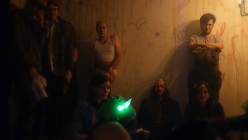 A dimly lit room with a group of people, some standing, some sitting, and a glowing green puppet in the foreground.