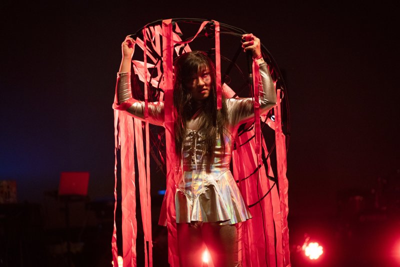 Performer in a silver costume holding a black circular frame with red fabric strips, illuminated by red stage lights.