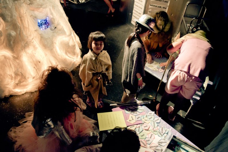 Children in costumes interact with a game and props in a dimly lit art installation.