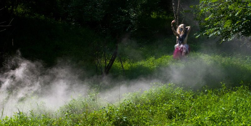 A person in a pink skirt and light top stands in a misty, sunlit green forest with arms raised.