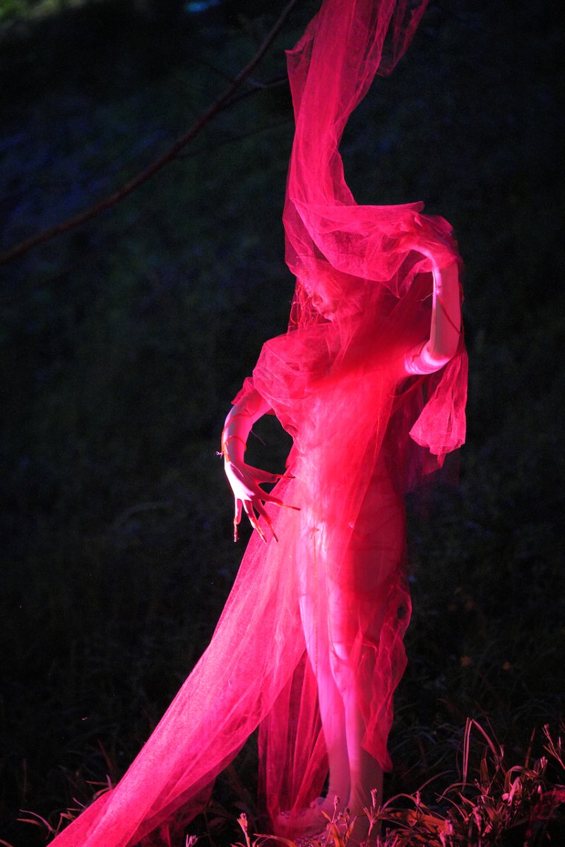 Figure shrouded in flowing bright pink fabric, arms raised, against a dark, blurred natural background.