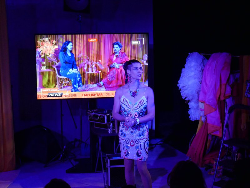 A person in a sequined dress stands before a large screen showing two women in traditional attire on a news broadcast.