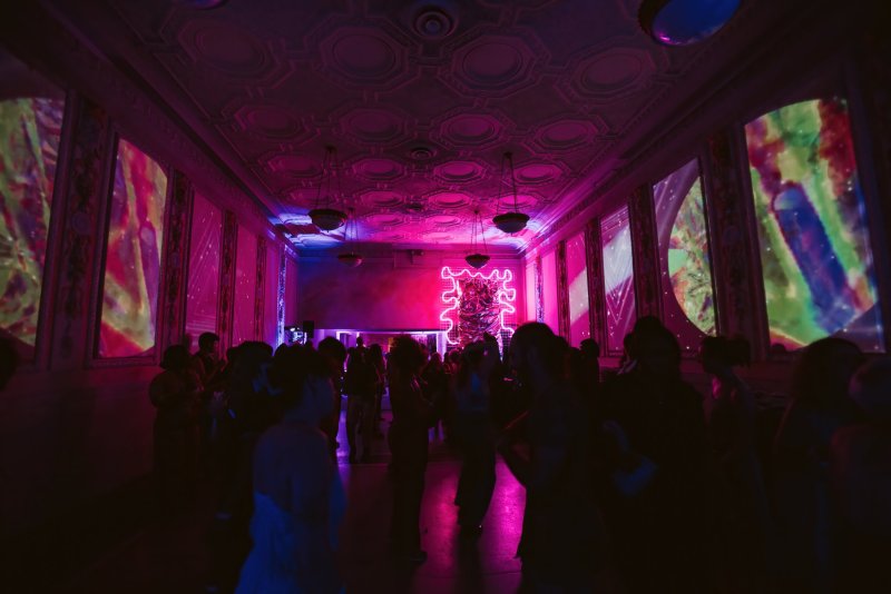 Dark room with silhouetted figures dancing under pink and purple lights, abstract projections on walls, and a glowing neon sculpture.