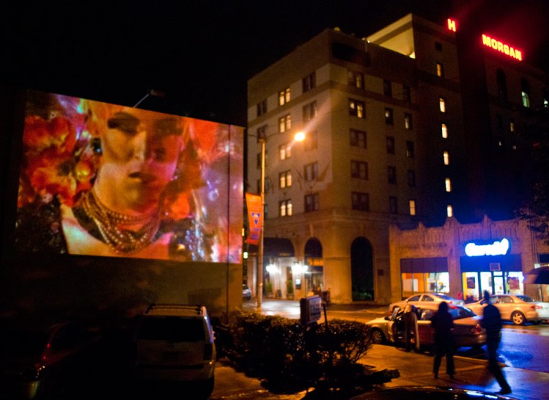 Nighttime street scene with a large projection of a golden-skinned figure adorned with jewelry and flowers on a building.