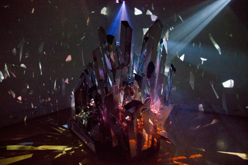 Large, jagged crystal sculpture reflecting blue, purple, and orange light in a dark room with scattered light shards.