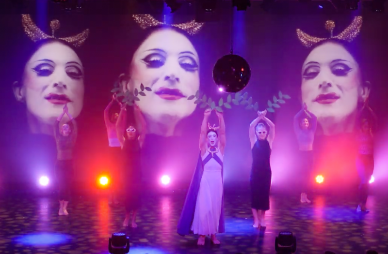 Performance with a central figure in a white dress and purple cape, flanked by dancers, under three large projected faces and a disco ball.