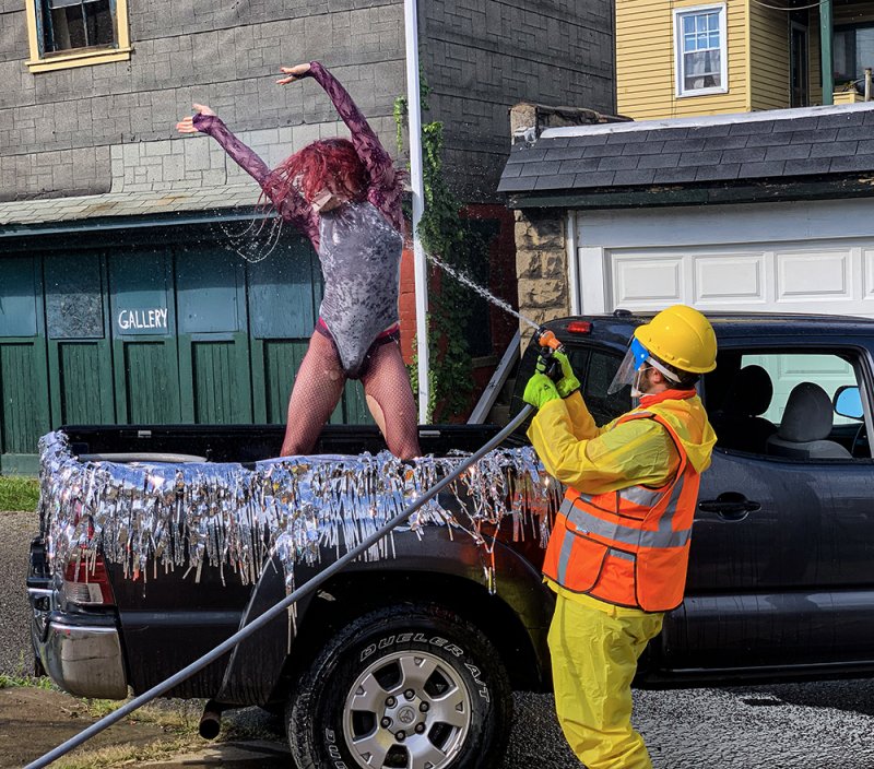 A person in a glittery bodysuit with red hair stands in a truck bed, being sprayed with water by a person in a hazmat suit.