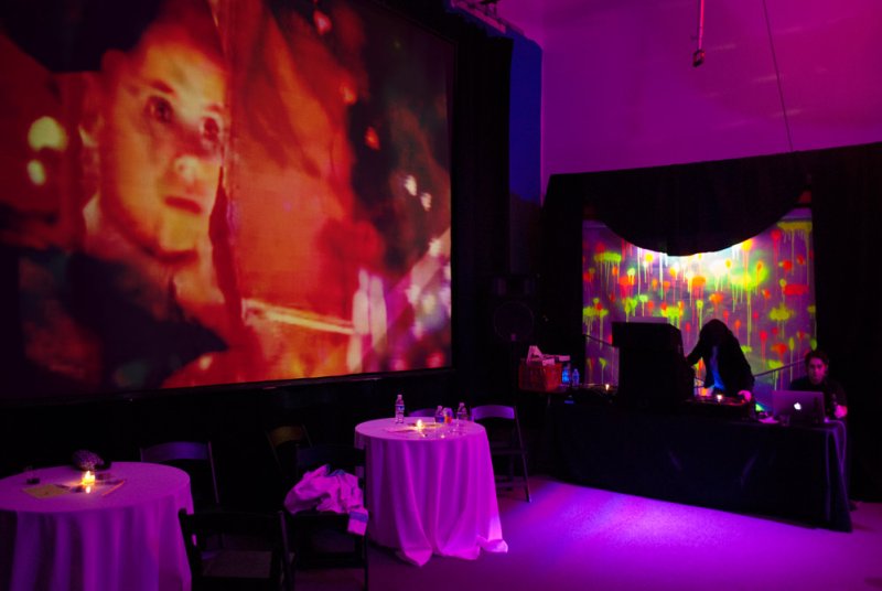 Installation view with a large video projection of a face and abstract shapes, tables with candles, and two people at a sound mixing station under purple lighting.