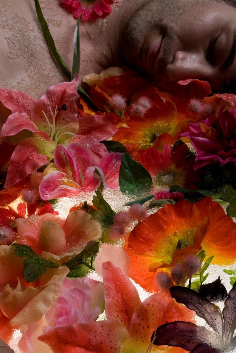 Close-up of a person's face partially submerged in water, surrounded by vibrant pink, orange, and red flowers.