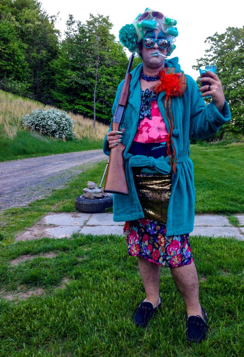 Person in teal robe, curlers, sunglasses, and floral dress holds a rifle and a drink outdoors.