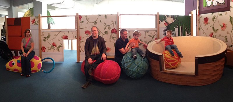 People sit on and around oversized yarn balls and a cat bed in a colorful, playful room.