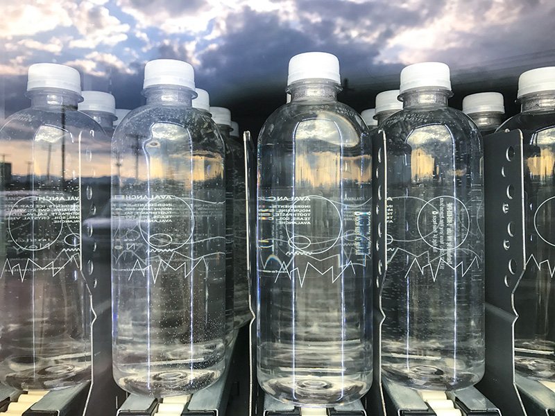 Row of clear plastic water bottles with white caps, some showing reflections of a cloudy sky, in a vending machine.