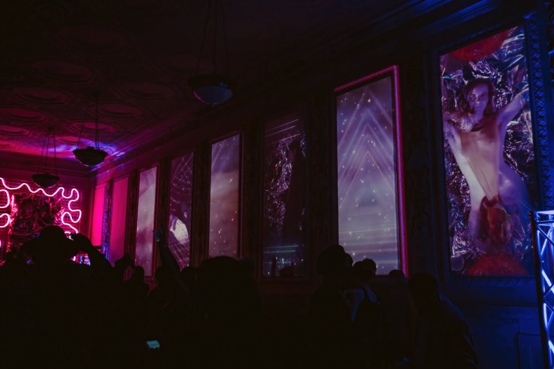 Dark room with people silhouetted against neon pink squiggles and purple-lit screens displaying abstract patterns and a nude figure.