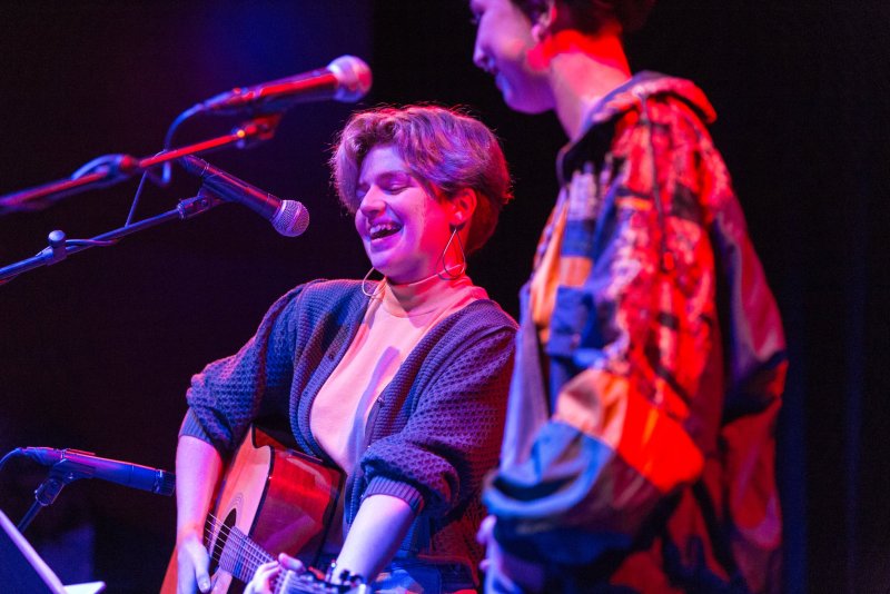 Musician with short hair smiles, playing acoustic guitar under red and blue stage lights. Second person stands to the right wearing a colorful jacket. Microphones visible.