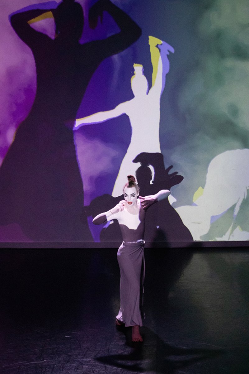 Performer in white with dramatic makeup and hair, dancing before a screen projecting large, colorful silhouettes.