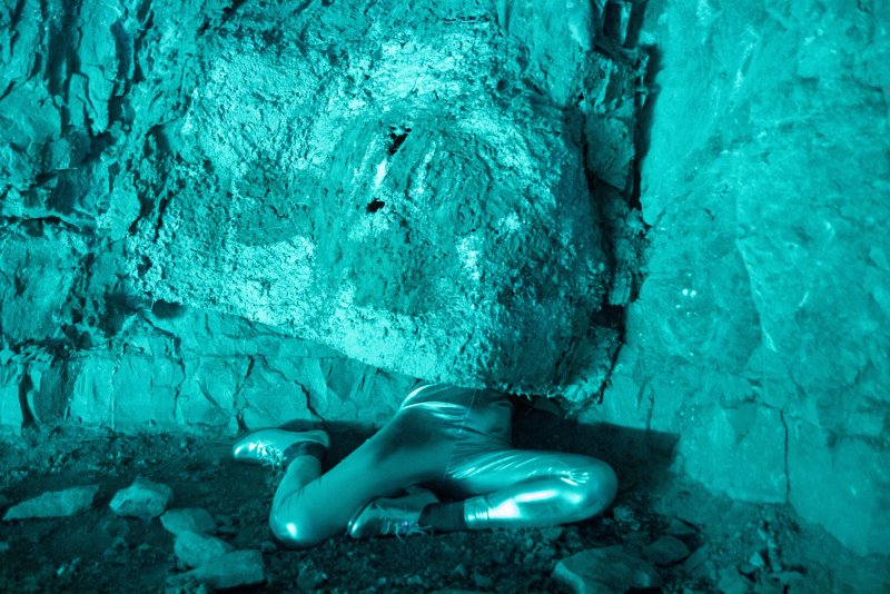 Person in shiny silver suit lying on dirt ground in a dark cave, illuminated by a bright teal light.
