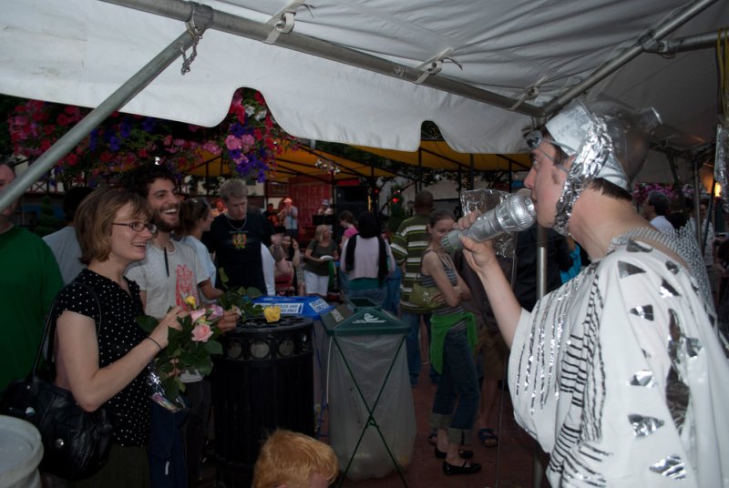 Man in silver costume sings into a microphone under a tent, surrounded by a diverse crowd and flowers.
