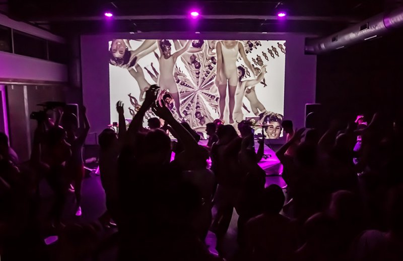 Crowd dancing in a dark room with purple lights, facing a screen displaying distorted, nude digital figures.