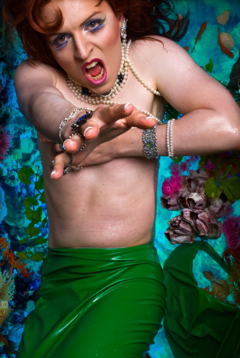 Person in a green skirt, pearls, and makeup, reaching forward with an open mouth, against a blue and coral reef background.