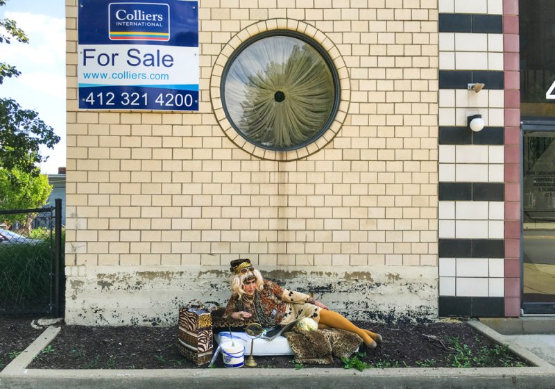 A woman in a patterned jacket and yellow tights reclines on a leopard print blanket before a beige brick wall.
