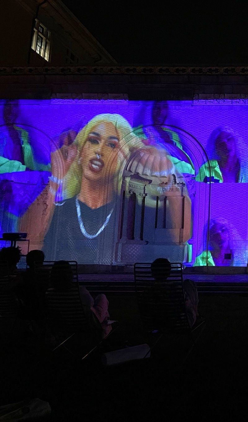 Drag queen with blonde hair, dark top, and silver chain projected on a building, with a dome structure in front and purple/green figures behind.