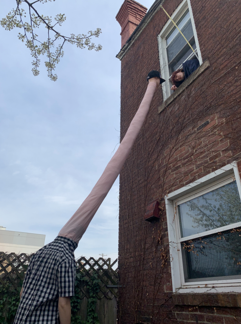 A person in a plaid shirt with an elongated neck reaching up to a window where another person looks out.